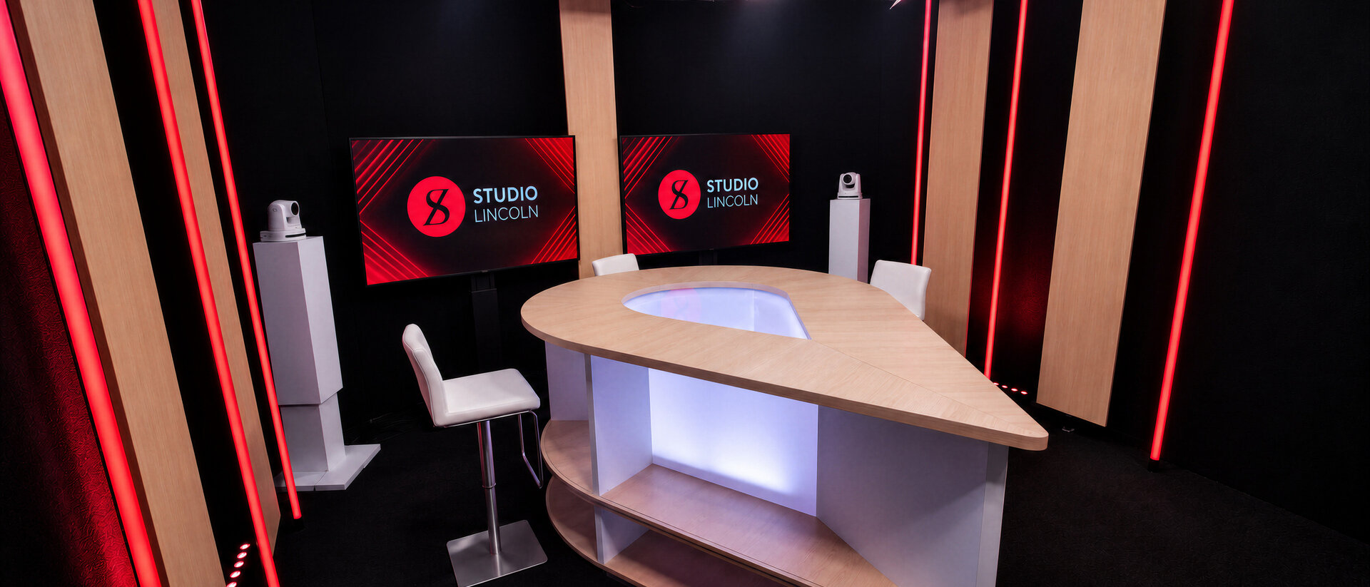 Le plateau en configuration table, un pupitre en bois éclairé, des tabourets blancs et deux écrans aux couleurs du studio