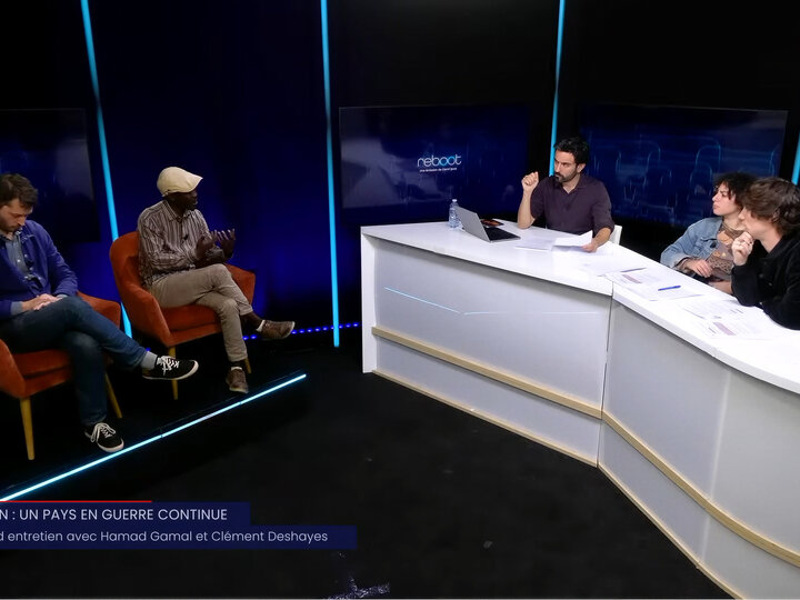 Un animateur et deux chroniqueurs à leur table, face à deux invités assis dans des fauteuils, sur un plateau éclairé en bleu