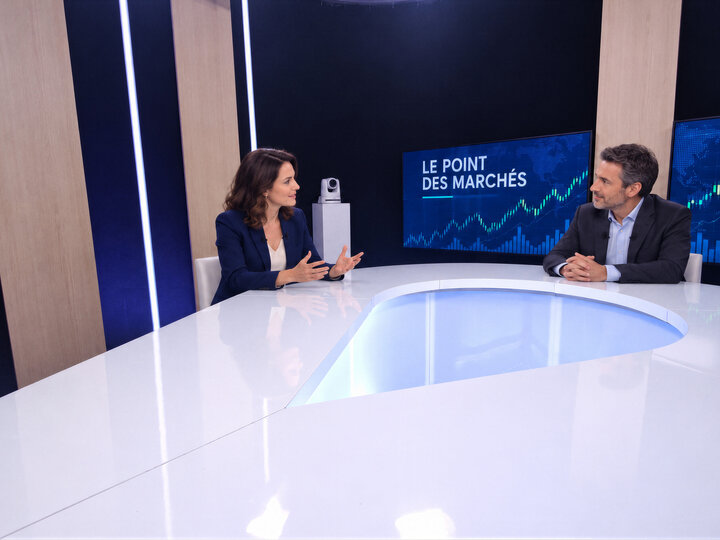 Une animatrice et un invité face à face à une table blanche, devant des écrans de cours de bourse